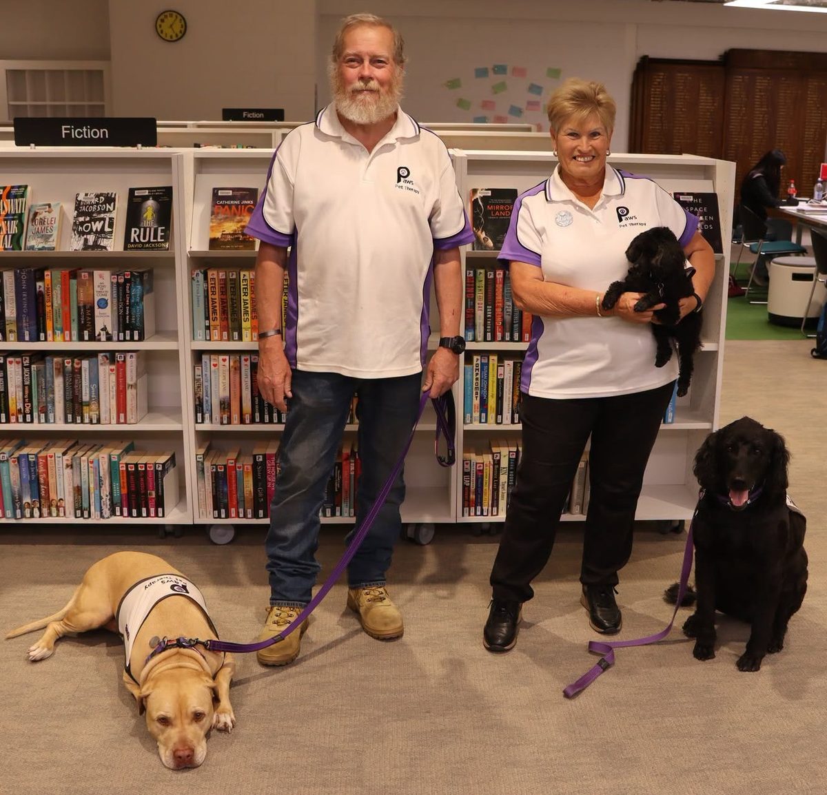 Two PAWS volunteers in a library with their dogs: a yellow labrador lying on the floor and two black dogs, one held in arms