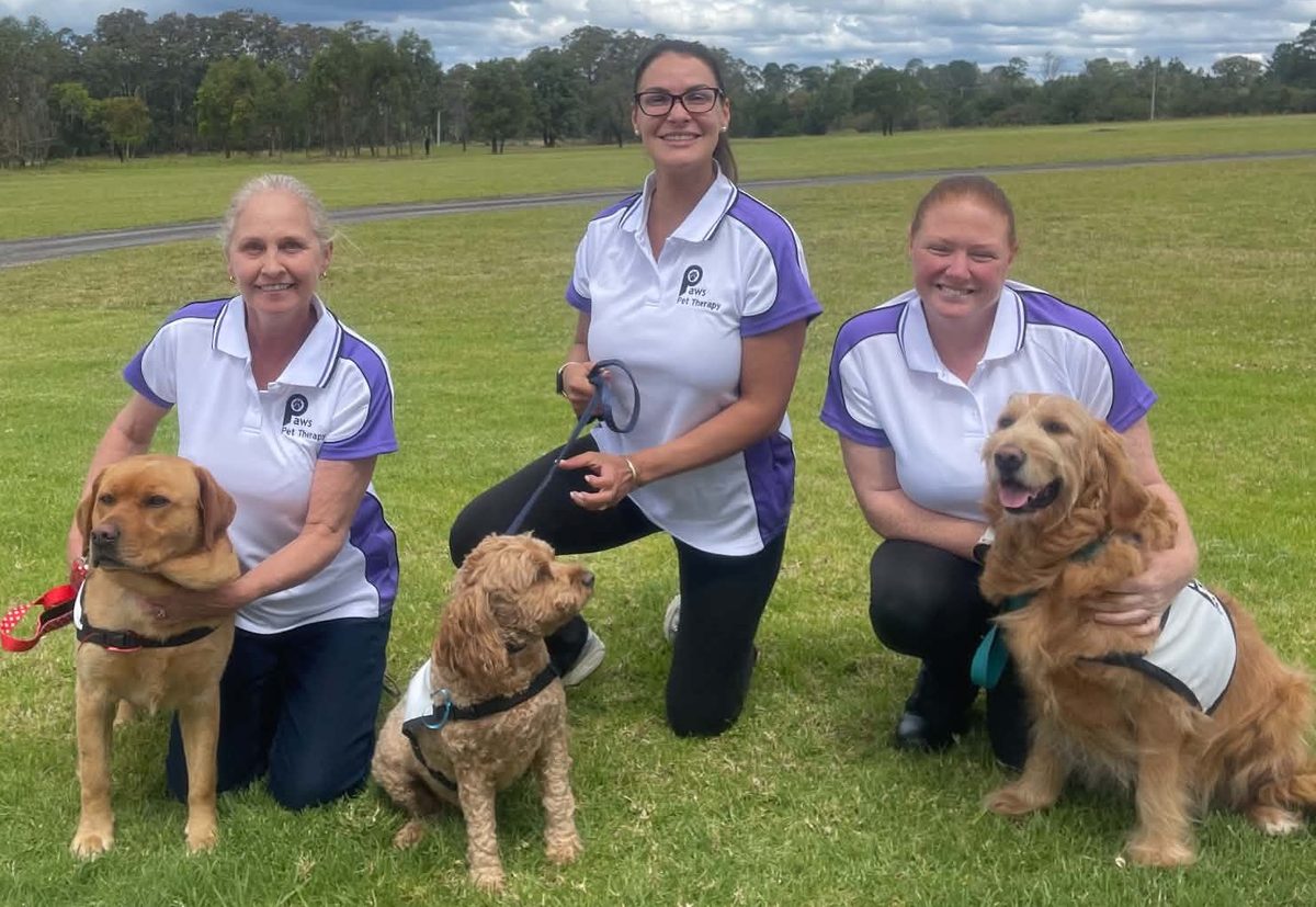 Three new PAWS volunteers in their PAWS shirts kneeling on the grass with their dogs at the Orangeville training day